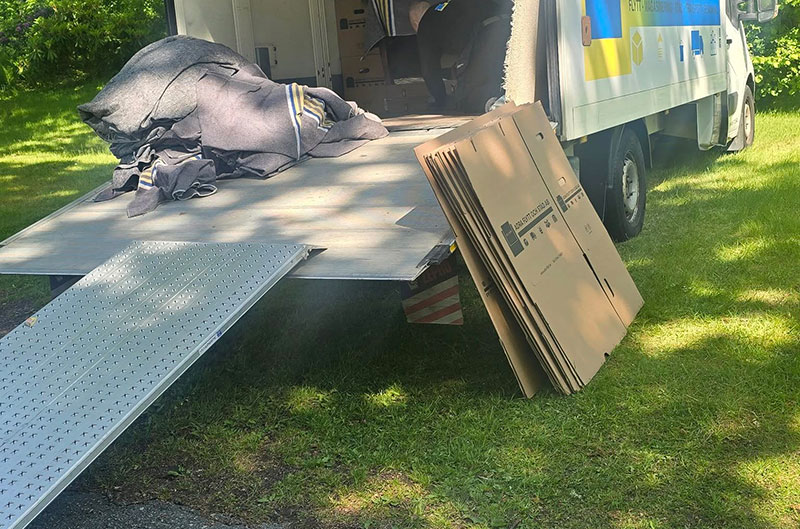 En flyttbil med öppen bakdörr står parkerad på en gräsmatta. Platta kartonger står staplade bredvid lastbilen och flyttfiltar ligger på lastbilens flak. Solljuset kastar skuggor på marken.