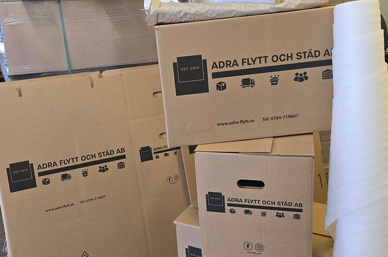 Flera flyttkartonger märkta "ADRA FLYTT OCH STÄD AB" är staplade på varandra, tillsammans med en rulle med packmaterial och delvis synliga inslagna föremål i bakgrunden.