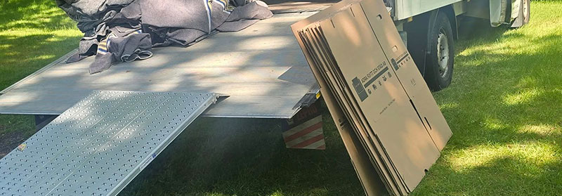 En flyttbil med öppet flak är parkerad på en gräsmatta. Flera hopvikta flyttfiltar ligger på flaket och en hög med stora kartonger står upprätt bredvid en metallramp.