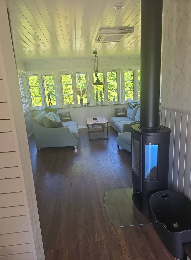 Ett ljust vardagsrum med trägolv, två ljusblå soffor, en vedeldad kamin, ett soffbord och stora fönster som släpper in naturligt ljus och utsikt över grönskan utanför.